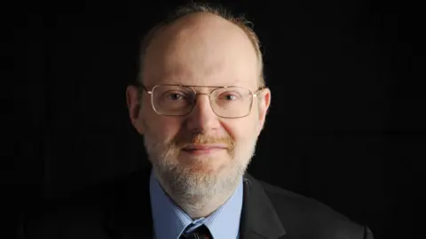 Martin Rosenbaum looks directly at the camera. He has a short beard with a mixture of brown, ginger and white hair and balding hair on his head. He has glasses which have a rounded frame and golden colour. He wears a formal shirt with a blue pinstripe, a mostly black, patterned tie and black suit. The background is dimmed so it's difficult to make out the location of the picture.