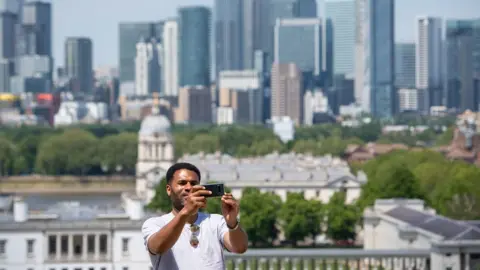 PA Media A man takes a selfie with the skyline of Canary Wharf, from the Royal Observatory in Greenwich Park, London