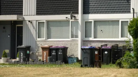 Ant Saddington/BBC Two terraced houses with four large rubbish bins and one small one lined up underneath their front windows.