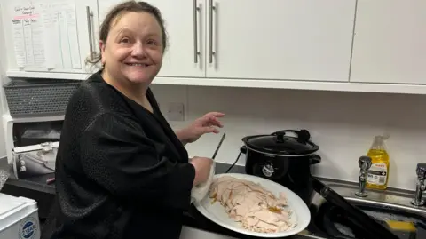 A woman in a black dress in a kitchen carving the turkey which is on a big white plate.