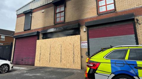 The exterior of the Welcome Organisation building with burn marks above the garage door which has been boarded up. A police car is also parked outside the building.