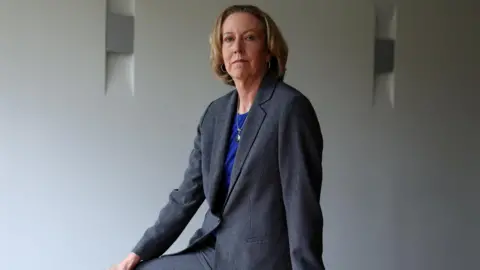 Reuters Newly appointed BP chief executive Meg O'Neill wears a grey suit and blue top while posing sat on the edge of a boardroom table.