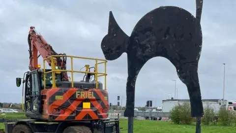 A sculpture of a black cat on the right, with its back arched and tail raised at the Black Cat roundabout, Cambridgeshire. On the left is a vehicle with an orange crane.