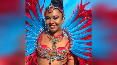 Sophia Estelle Mangroo Sophia Estelle Mangroo in a red and blue carnival outfit. It includes a vibrant spread of feathers behind her back, and an intricately beaded bra-style top, with a jewelled necklace and headpiece to match. She also has red and blue eyeshadow on, and red lipstick. Her hair is in a bun on top of her head. 