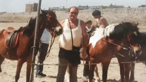 Jay Bateman A man in a white vest is stood between horses on a sandy beach.