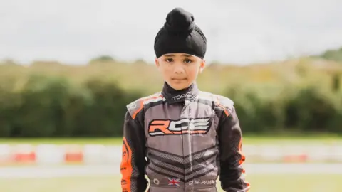 Monkey Media Productions Dhian Singh standing with his grey orange and black racing suit on and holding his helmet in his left hand.