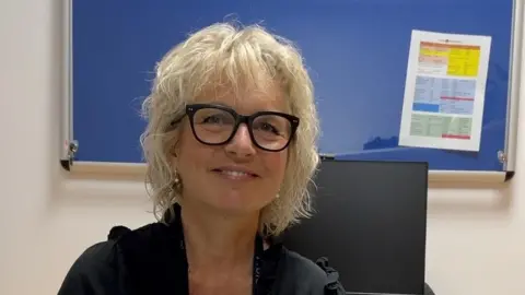 Carroll Weston/BBC Woman with light curly hair and glasses sits in front of a noticeboard