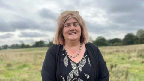 Kate Bradbrook/BBC Women with medium-length brown hair and wearing a blue sweater stands in a field