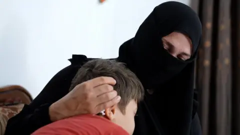 BBC Alia is wearing a black niqab and is embracing her son. He is wearing a red t-shirt and has brown short hair.