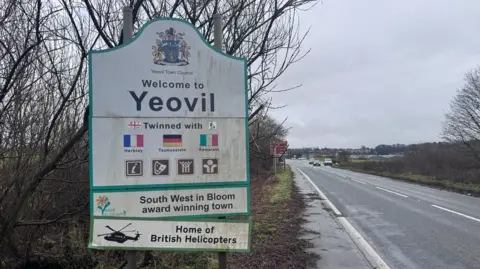 A road sign reading 'welcome to Yeovil' with a further sign lower down which is partly attached and slightly hanging off reading 'Home of British Helicopters'