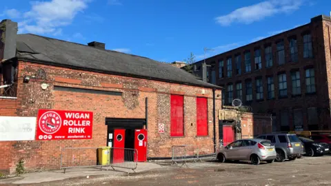 The outside of the roller rink - a red-brick building with large red doors and a red sign reading 'Wigan Roller Rink'.