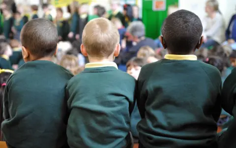 BBC The image shows three children sitting side by side, viewed from behind. They are wearing matching dark green sweaters with yellow collars, suggesting a school uniform. The background is slightly out of focus, but it looks like an indoor space with bright colours, including a green wall or partition.