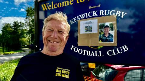 Alan Dunn standing beside a big sign at the entrance to the rugby ground