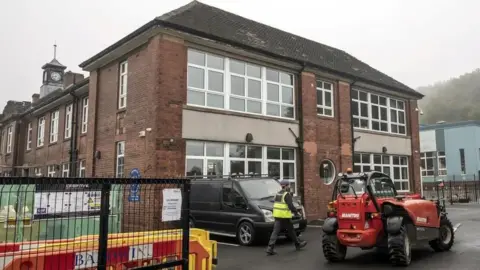 A workman and machinery is outside a brick built school building