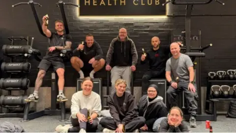 Josh Baines A group of people sat surrounded by gym equipment and in front of a sign that says The Atrium Health Club.