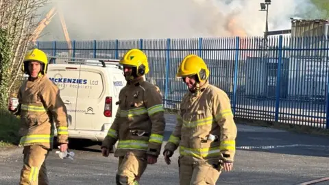 Three firefighters wearing full protective gear and bright yellow helmets walk along a road near an industrial site. Fire hoses run along the ground beside them, and thick smoke is rising in the background behind a metal fence.