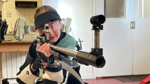 Shooter Libby Hart aims the gun down range wearing protective gear which include gloves, jacket, a hat which blokes out her right eye. 