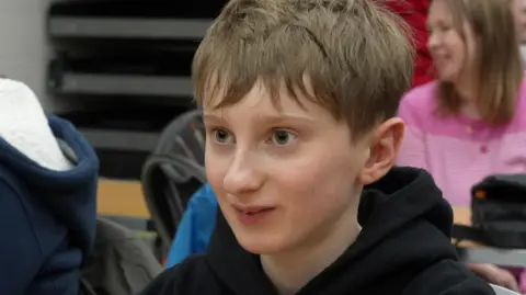 Taylor, a young boy with light brown hair, looking to the side of the camera. He is smiling and wearing a black hoody jumper.
