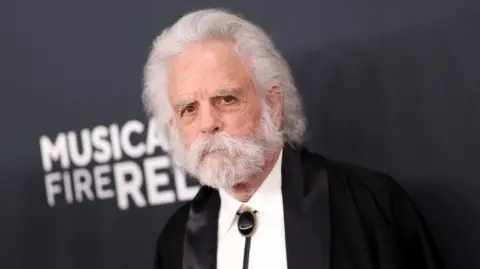 Bob Weir, with a dark suit and shite shirt, has a white beard and moustache