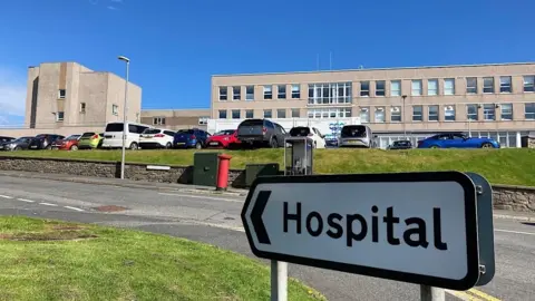 A sign pointing to a hospital. There's a large brown building in the background, with a lot of cars parked in front of it