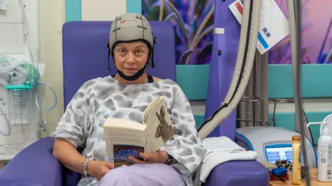 A woman in a hospital chair in a hospital ward wearing a scalp cooling machine on her head which is grey with a black chin strap. She is wearing a grey and white blouse and reading a book.