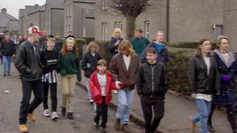 A protest photo at Raploch in the 1990s. Many local people walk on the street with houses in the background.