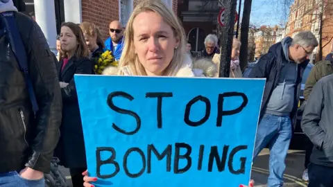 Elena Shevchenko holds a sign reading "Stop bombing my family"