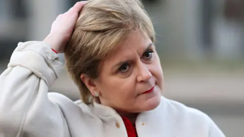 Nicola Sturgeon standing outside, wearing a white coat and a red top. Her right arm is raised and she is touching her hair, which is short and sandy blonde. Her expression is neutral.