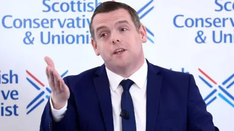 A man with dark, wearing a dark blue suit and tie, speaks with his right hand raised in front of him. He is standing in front of a Scottish Conservative-branded background  