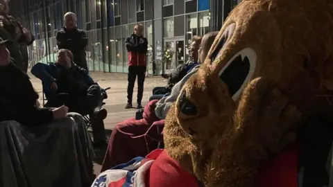 Supplied Someone wearing a large lion-head costume sitting on a camping chair outside the LNER Community Stadium at night.