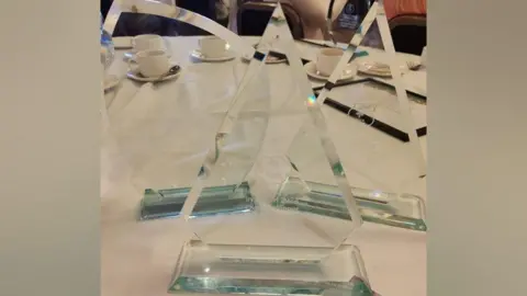 Shrewsbury Town Council Three glass awards sitting together on a table, with teacups and saucers in the background