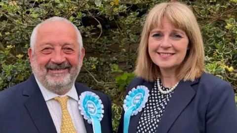 A man and a woman are wearing light blue and white rosettes featuring the Reform UK logo. The man, on the left with grey hair and a beard, and the woman, with blonde/brown hair, are both wearing blue jackets.