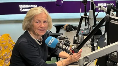 A woman with short blonde hair and a pearl necklace sitting at a radio microphone in a studio