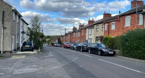 BBC A view of Midland Terrace in Westhouses, Derbyshire. Several cars can be seen parked either side of the road with terraced houses on both sides 