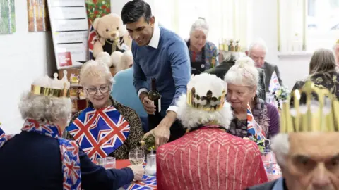 PA Media Prime Minister Rishi Sunak pours a drink for a member of a community group's lunch club at Mill End Community Centre, Rickmansworth, as part of the Big Help Out, to mark the crowning of King Charles III and Queen Camilla