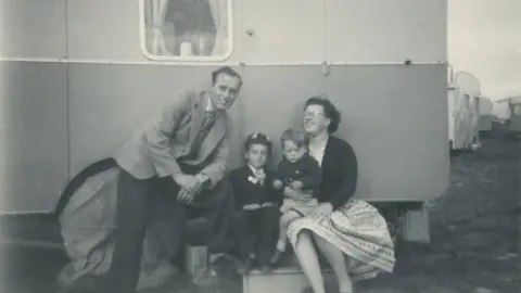 Roy Daines Black and white photo of mother, father and two sons (one wearing a cap) outside a caravan