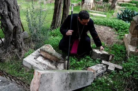 Reuters Archbishop Hosam Naoum inspects a desecrated grave at the Protestant Cemetery on Mount Zion in Jerusalem (4 January 2023)