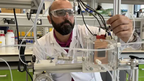 BBC Prof Enrico Andreoli in a lab wearing a lab coat and safety glasses looking at an experiment