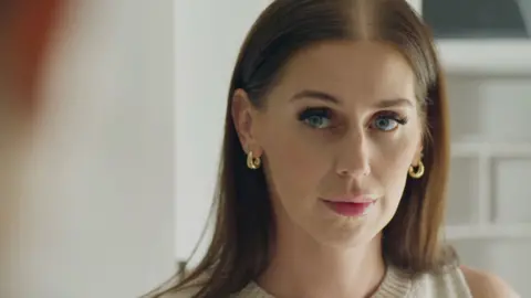 Jen, a woman with dark hair, looks to the presenter. She wears make-up and gold hoops.