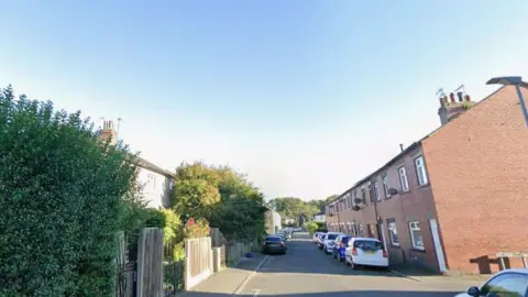 Google Dale Street in Ulverston. A straight, narrow road with different styles of two-storey terraced houses. On the left there are fences, walls and large bushes and trees separating houses from the narrow pavement which parked cars straddle further down. On the right, red brick houses open directly on to the pavement. Cars are also parked all along that side but are fully on the road rather than part on the pavement.