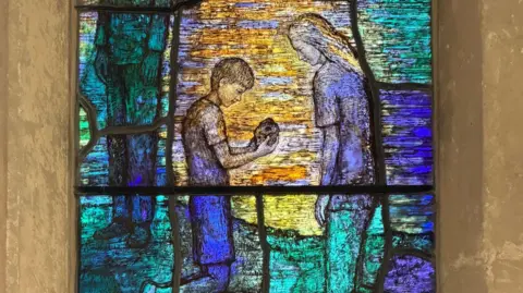 Image of a stained glass window depicting a boy who is kneeling down looking at a shell. A young girl is standing over him while a second girl looks on.