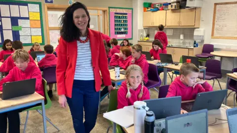 Clare Brodie standing in a classroom next to a girl with pigtails who is sitting at a desk with a notebook and a laptop, and is looking at the camera. There are more than a dozen pupils sitting at desks, all wearing deep pink hoodies. Ms Brodie is wearing a red jacket, white striped top and jeans.