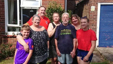 Rhian Collinson A family group photo of seven people of varying ages. It is a sunny day and they are stood outside on grass, in front of a house, all smiling at the camera.