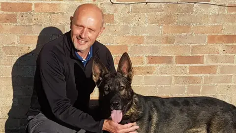 Ian Partington kneeling, holding a dark brown German Shepherd, which has its tongue out. Ian, who is bald, is wearing all black and smiling. They are in front of a brick wall.
