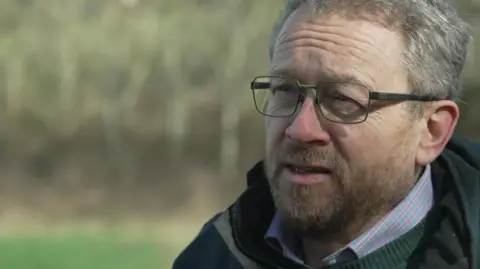 Green councillor Chris Copeman, how is wearing a pink and blue shirt with a green jumper, coat and glasses, is standing in a field as he is being interviewed. 