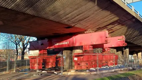 A temporary prop underneath the Gateshead Flyover. It is a huge red metal structure and is fenced off. The flyover above is made from concrete.