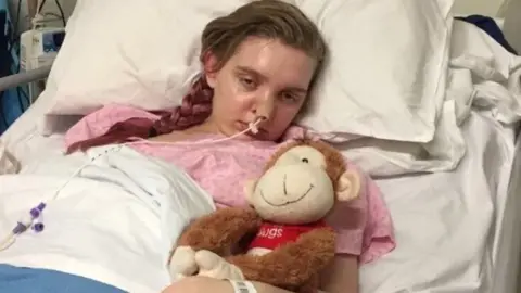 Lucy Dawson Lucy lies in a hospital bed and clutches a stuffed toy. Her eyes are unfocussed and she looks pale and poorly