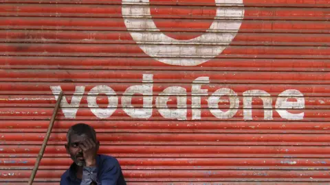 Getty Images A man sits outside the downed shutters of a shop painted with a logo of Vodafone on its shutter in Mumbai, India on 24 February 2019