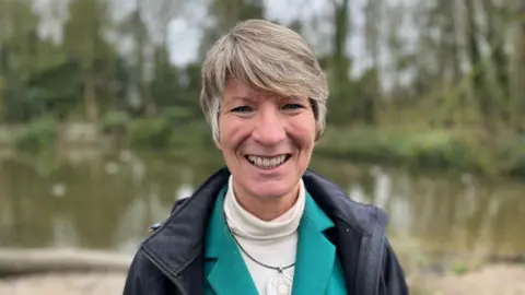 Tom Jackson/BBC Pippa Heylings stands by a river. She is smiling and wears a waterproof coat with a teal jacket and white jersey underneath.