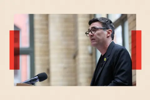 Reuters Andy Burnham delivers a speech at the People's History Museum in Manchester. He is viewed in profile standing at a podium. In the background are cream tiles and windows with green frames. Burnham has dark greying hair and wears dark-rimmed glasses and a dark jacket with a bee logo pin badge on his lapel.